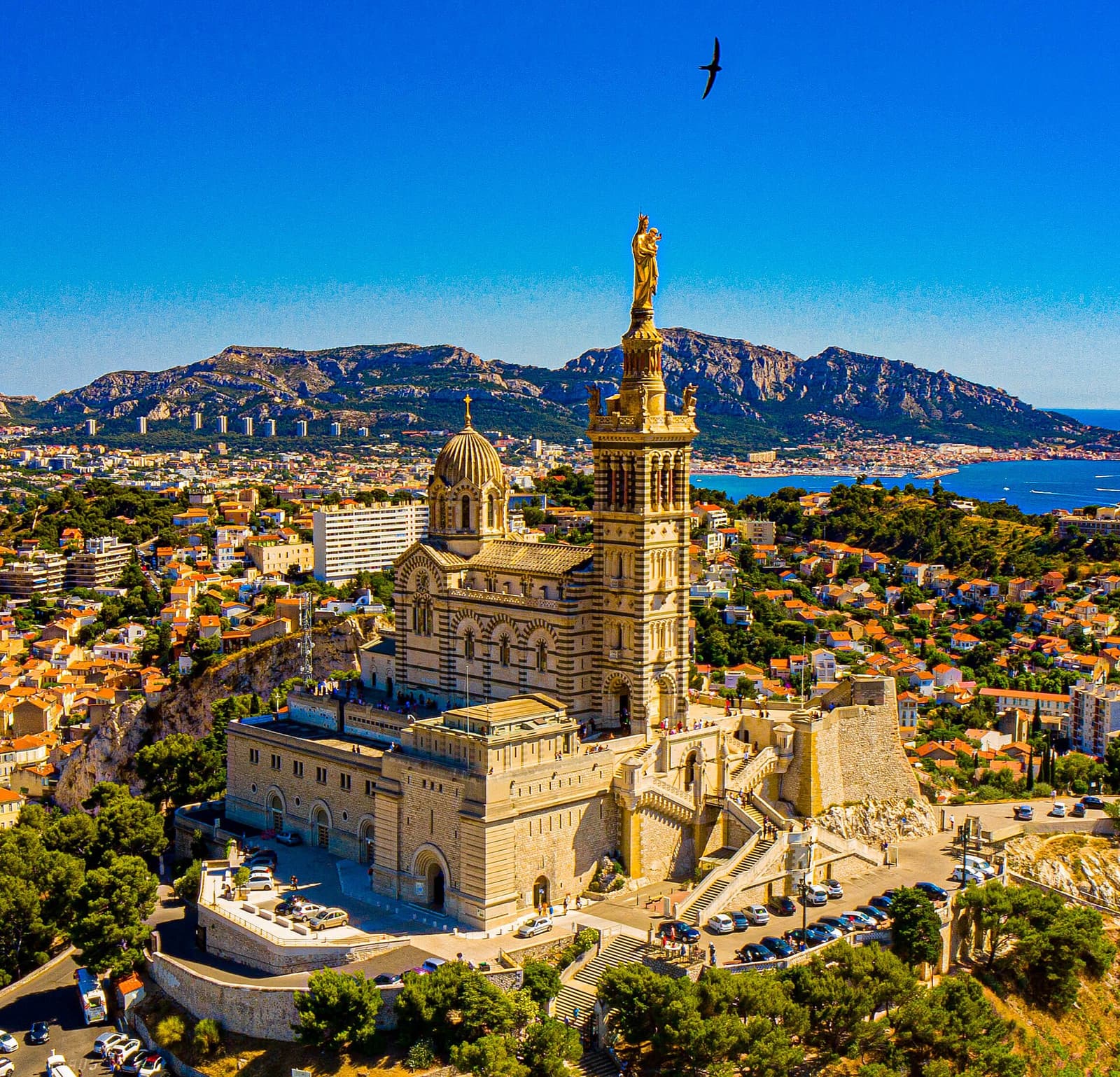 Luftbild der Basilika Notre-Dame de la Garde auf ihrem Hügel mit Marseille und den Bergen dahinter