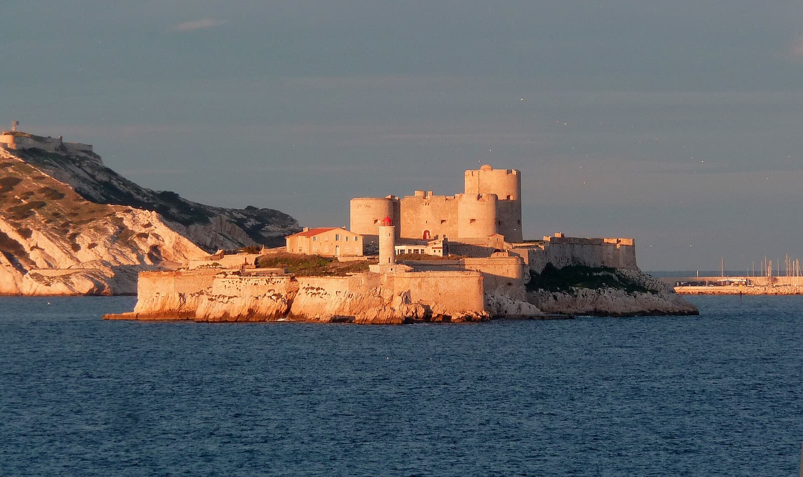 The Château d'If glowing in warm low sunlight with dark cliffs behind