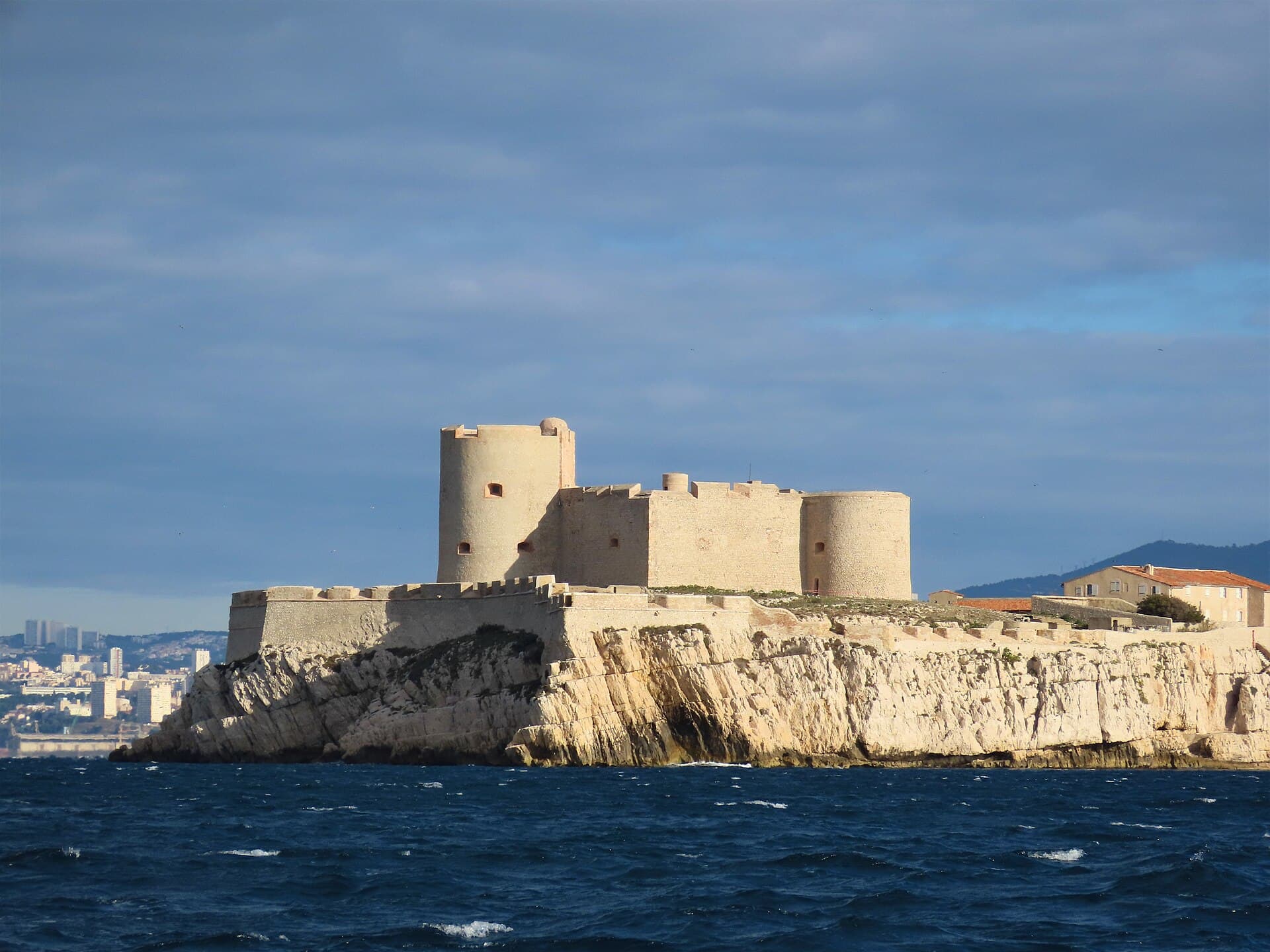 Das Château d’If, eine helle Steinfestung mit Rundtürmen auf ihrer Felseninsel, unter blauem Himmel mit Marseille in der Ferne